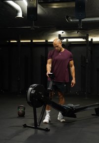 Άνδρας με πουκάμισο βουργουνδί προσαρμόζει τον πίνακα ελέγχου μηχανής κωπηλασίας σε γυμναστήριο με χαμηλό φωτισμό, με kettlebell στο πάτωμα κοντά του.