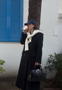 Γυναίκα με μαύρο παλτό και μπλε καπέλο πίνει καφέ, κρατώντας μαύρη τσάντα, στέκεται δίπλα σε λευκό τοίχο με μπλε παντζούρια παραθύρου και κορμό δέντρου.