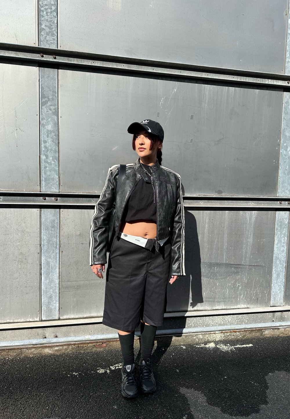 Μαύρο δερμάτινο μπουφάν μηχανής με λευκές ρίγες, μαύρο κοντό τοπ, μαύρες σορτς, μαύρες κάλτσες και μαύρα αθλητικά παπούτσια; στέκεται μπροστά σε ένα μεταλλικό φόντο.