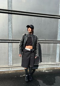 Μαύρο δερμάτινο μπουφάν μηχανής με λευκές ρίγες, μαύρο κοντό τοπ, μαύρες σορτς, μαύρες κάλτσες και μαύρα αθλητικά παπούτσια; στέκεται μπροστά σε ένα μεταλλικό φόντο.