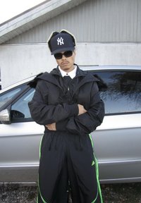 Casaco bomber preto com capuz forrado de pelo, combinado com calças pretas a condizer com riscas verdes. Acessórios incluem um boné preto e óculos de sol.