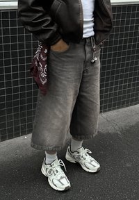 Brązowa kurtka bomberka ze skóry, szare szerokie szorty dżinsowe, białe skarpetki oraz jasnoszare sneakersy z ciemnymi akcentami. Czerwona bandana w kieszeni.