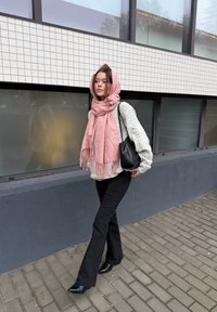 Világos rózsaszín rojtos sál, krém színű strukturált pulóver, fekete bővülő szárú nadrág és hegyes orrú fekete csizma. Városi környezet, csempézett fal mellett.