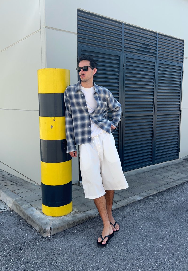Homme portant des lunettes de soleil, une chemise à carreaux, un short blanc et des tongs, appuyé contre un poteau rayé jaune et noir près d'un bâtiment.