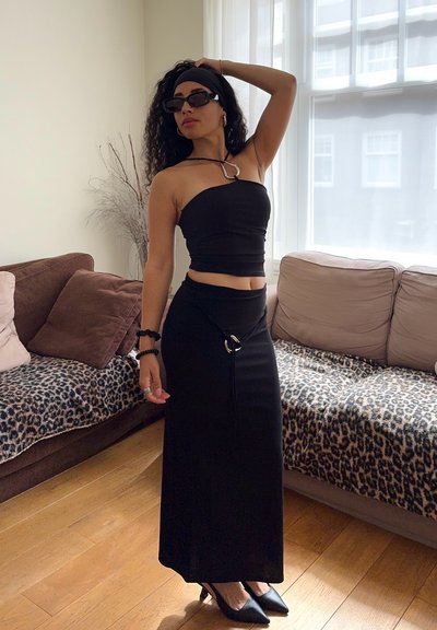 Top bandeau negro combinado con una falda larga negra, que presenta un acento metálico y cordones, complementado con elegantes tacones negros y gafas de sol.