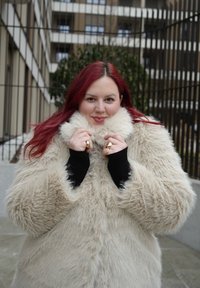 Γυναίκα με μακριά κόκκινα μαλλιά που φοράει μεγάλο μπεζ παλτό από συνθετική γούνα, κρατώντας τον γιακά κοντά στο πρόσωπό της, στέκεται έξω δίπλα σε κτίρια και κιγκλίδωμα.