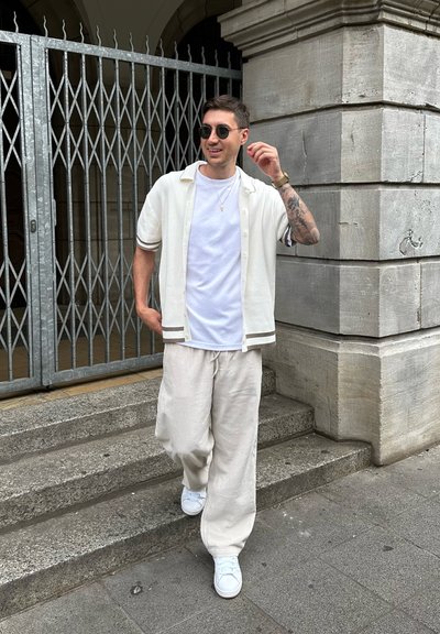 Camisa corta de color beige claro con detalles marrones, camiseta blanca, pantalones amplios a rayas claras y zapatillas blancas, contra una pared de piedra.