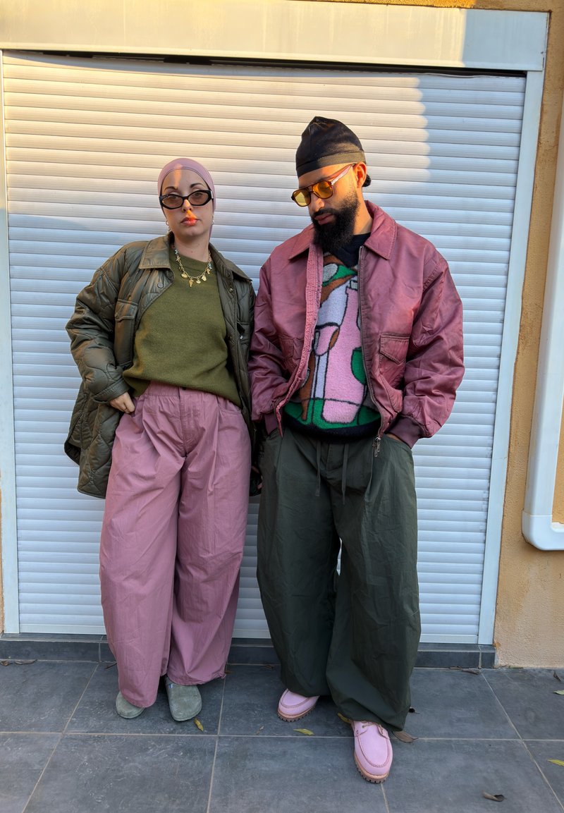 Deux personnes se tiennent devant un volet blanc. L'une porte une doudoune verte, un pull vert et un pantalon rose ; l'autre porte une veste bomber bordeaux, un pull à motifs et un pantalon olive.