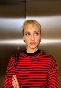 Jeune personne aux cheveux blonds courts portant des boucles d'oreilles argentées en forme d'anneau et une chemise rayée rouge et noire, regardant sur le côté avec les bras croisés.