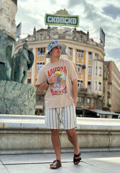 Camiseta gráfica beige con el texto "ARIZONA", pantalones cortos a rayas en azul y blanco, sandalias marrones y un sombrero de cubo con un patrón azul. Fondo urbano.