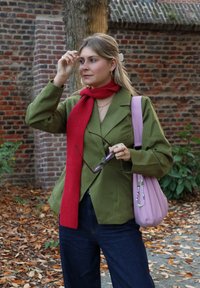 Olivengrønn blazer med tynne striper, rød ribbestrikket skjerf, mørke denimbukser, holder briller, rosa skulderveske med dekorative perler, bakgrunn av høstløv.