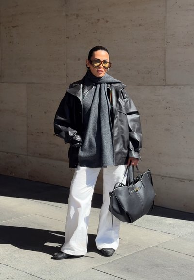 Mujer con chaqueta de cuero negra de gran tamaño, bufanda gris, pantalones blancos, gafas de sol, sosteniendo un gran bolso negro, de pie contra una pared de piedra.