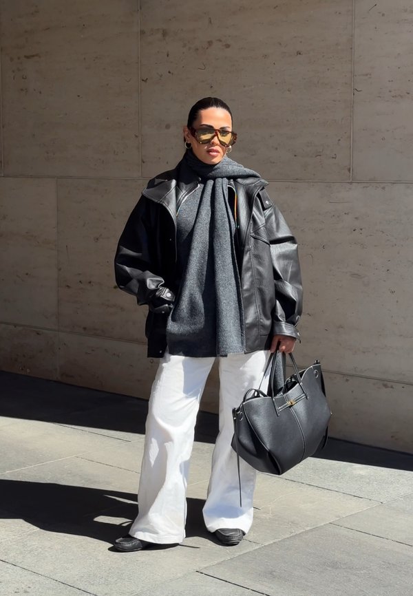 Femme portant une veste en cuir noire surdimensionnée, une écharpe grise, un pantalon blanc, des lunettes de soleil, tenant un grand sac à main noir, debout contre un mur en pierre.