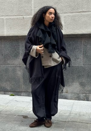 Mujer con cabello rizado que lleva un chal negro, chaqueta de cuadros cruzada, pantalones negros sueltos y zapatos marrones, de pie en la acera de una ciudad.