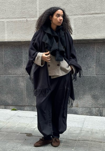 Mujer con cabello rizado que lleva un chal negro, chaqueta de cuadros cruzada, pantalones negros sueltos y zapatos marrones, de pie en la acera de una ciudad.