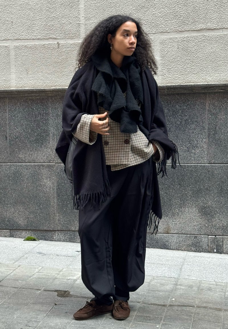 Femme aux cheveux bouclés portant un châle noir, une veste croisée à carreaux, un pantalon noir ample et des chaussures marron, debout sur un trottoir en ville.