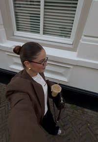 Brun oversized blazer over en hvit skjorte, svarte bukser, holder en klar kopp med lokk og sugerør. Fyrkantede briller og gulløredobber.