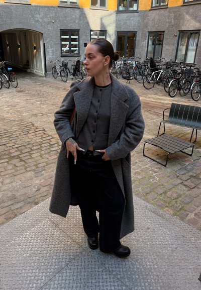 Joven con el cabello peinado hacia atrás de pie en un patio, lleva puesto un abrigo largo gris, un cárdigan abotonado y pantalones anchos negros, con bicicletas al fondo.