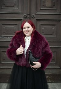 Μια γυναίκα που χαμογελάει, φορώντας ένα μπορντό τεχνητό γουνάκι και μαύρη φούστα, στέκεται μπροστά σε μεγάλες ξύλινες πόρτες κρατώντας μια μαύρη τσάντα.