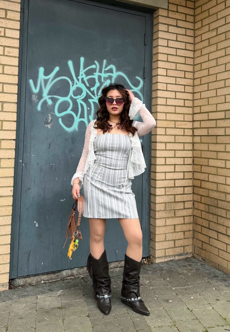 Femme en robe rayée, cardigan en dentelle, lunettes de soleil en forme de cœur, tenant un sac à main, debout devant une porte bleue couverte de graffitis.