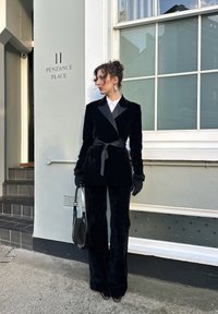 Blazer en velours noir avec revers en satin, pantalon assorti et gants. Un col blanc apporte un contraste. Les accessoires comprennent un sac à main noir.