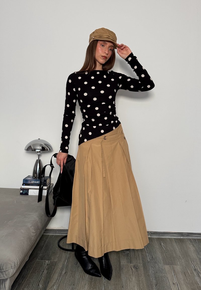 Femme portant une casquette beige, un haut noir à pois, une jupe beige et des bottes noires, tenant un sac noir, debout à côté d'un canapé gris et d'une lampe.