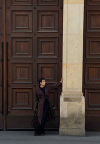 A person in a dark trench coat is leaning against a beige stone pillar, holding a large bag, in front of a large wooden door.