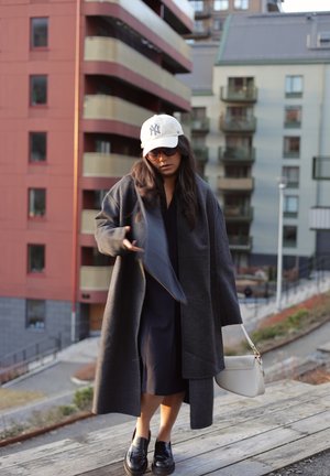 Abrigo largo y oscuro sobre un vestido negro, zapatos de charol negros, bolso beige, gorra beige y gafas de sol en un entorno urbano con edificios modernos.