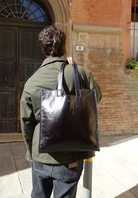 Black leather tote bag with a smooth texture, twin handles, and minimalist design. Carried on the shoulder by someone wearing an olive green jacket.