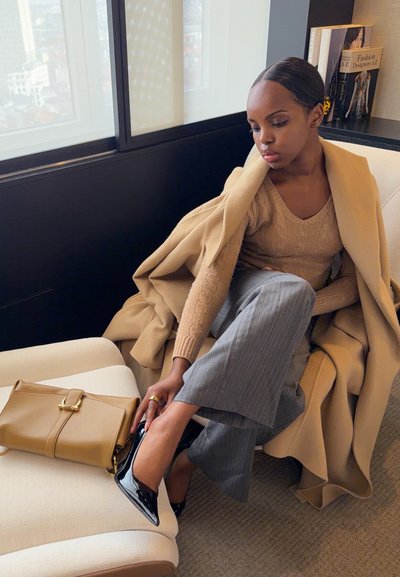 Mujer sentada junto a una ventana, ajustándose un zapato de tacón alto de charol negro, vestida con un abrigo color camello, suéter beige, pantalones grises, con un bolso beige cerca.