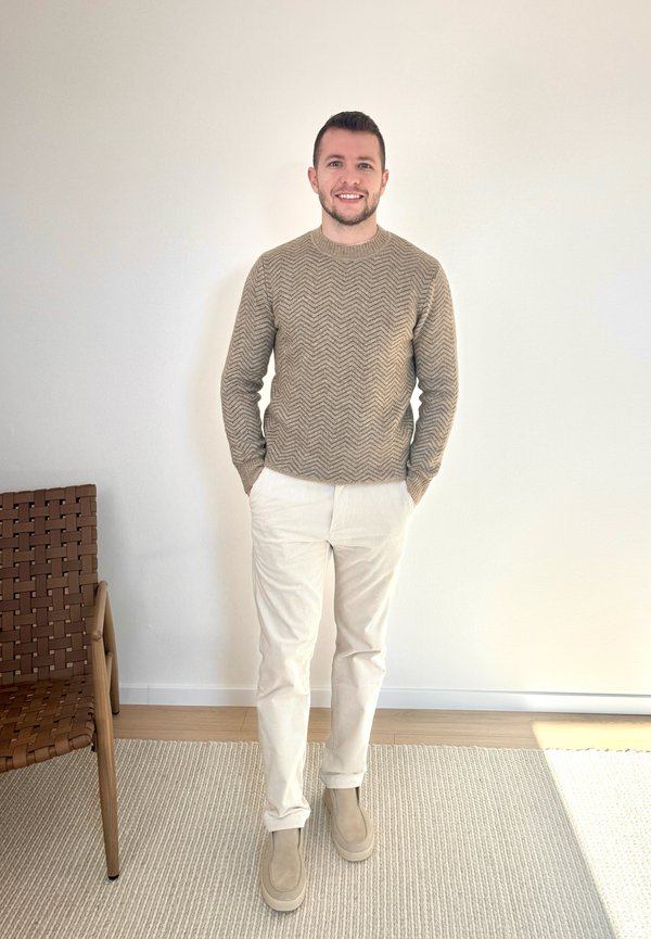 Moški se smeji, stoji v notranjosti, oblečen v bež pulover s vzorcem, bele hlače in bež čevlje poleg tkanega stola na svetli preprogi.