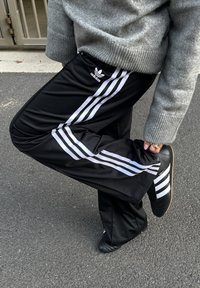 Fekete melegítőnadrág három fehér csíkkal, rugalmas derékrésszel és apró Adidas logóval. Fekete cipővel párosítva.