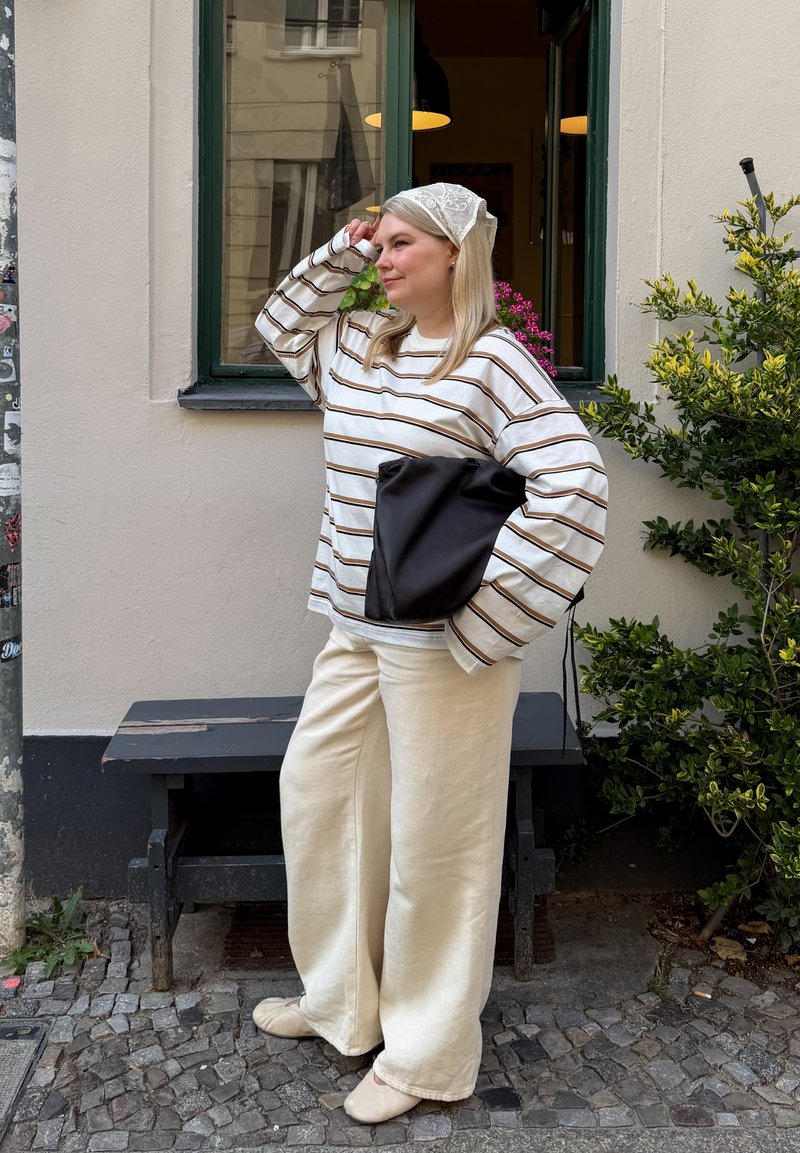 Femme portant une chemise rayée à manches longues et un pantalon crème, debout dans une rue pavée tenant un sac noir près d'une fenêtre avec des plantes.