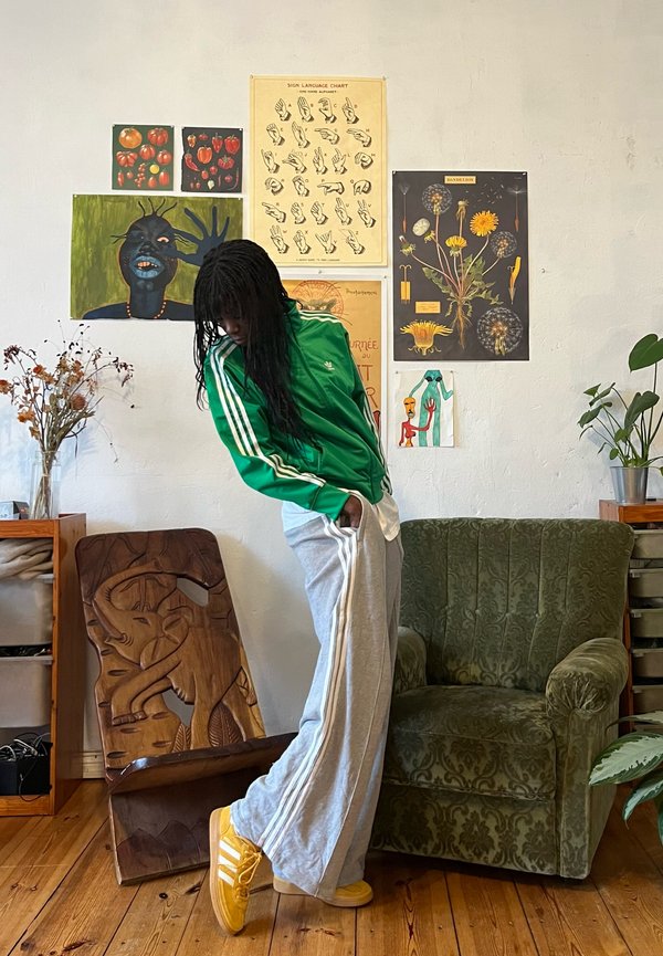 Groene ritsjack met witte strepen, grijze sweatpants en gele sneakers. Houten houtbewerking en groene beklede stoel op de achtergrond.