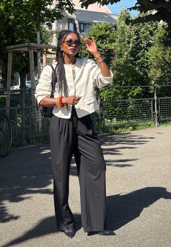Light-coloured cropped jacket, black wide-leg trousers, pointed black shoes, and accessories including a multicoloured bracelet and sunglasses.