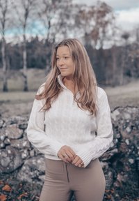 Hvit kabelstrikket genser med kvartglidelås, med ribbestrikkede mansjetter, båret over beige tettsittende bukser, satt mot en naturlig steinbakgrunn.