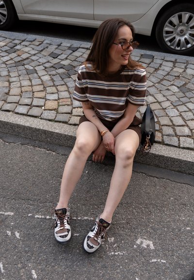 Camiseta a rayas marrón y blanca, pantalones cortos marrones y zapatillas distintivas en blanco y negro con diseño recortado. Los accesorios incluyen gafas de sol y un bolso.