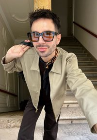 Beige mikina na zip, tmavé tričko, hnědé kalhoty. Tlusté černé sluneční brýle s oranžovými skly. Hnědý vzorovaný šátek uvázaný kolem krku.