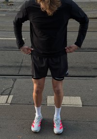 Személy fekete hosszú ujjú ingben, fekete rövidnadrágban, fehér zokniban és szürke sportcipőben, élénk rózsaszín csíkokkal, villamospályás utcán áll.