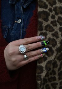 Montre à anneau en argent sur une main avec de longs ongles colorés présentant divers motifs. L'arrière-plan montre un tissu à imprimé léopard et du denim.