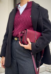 Bolso bandolera de cuero burdeos con un diseño sencillo, correa negra y herrajes dorados. Combinado con un suéter de punto burdeos y un abrigo negro.