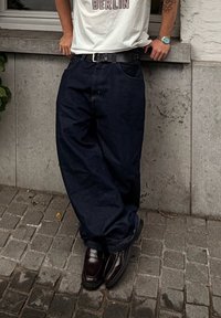 Osoba se oslanja na betonski zid, nosi tamne široke traperice, crni pojas, bijelu majicu s natpisom "BERLIN" i tamne sjajne cipele.