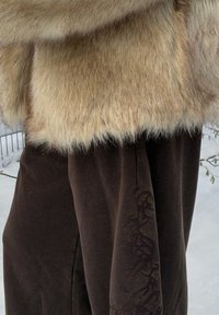 Manteau en fausse fourrure de couleur crème claire, présentant une texture dense. Pantalon marron avec un motif sombre le long du côté, complétant la tenue.
