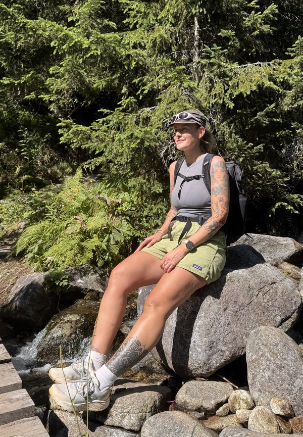 Jeune femme tatouée en tenue de randonnée assise sur un rocher près d’un ruisseau de forêt par une journée ensoleillée.