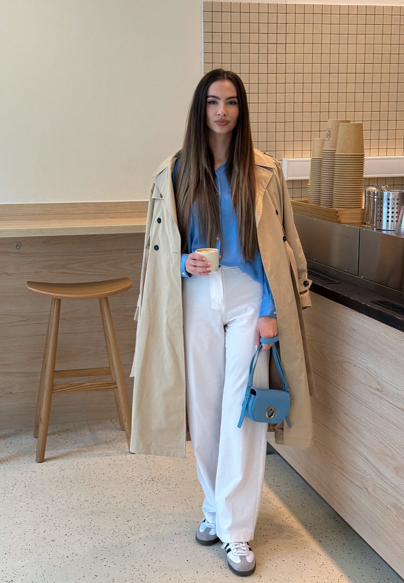 Femme en trench beige, blouse bleue et pantalon blanc tenant une tasse de café et un sac à main bleu dans l’intérieur d’un café moderne.