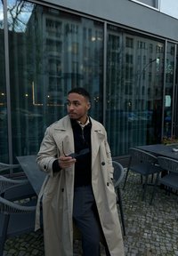 Kermankeltainen trench coat, jossa on suuret napit, mustan neuleen päällä, pitäen aurinkolaseja. Raidalliset harmaat housut. Ulkoilmakahvilan ympäristö, jossa on lasi- ja kivirakenteita.