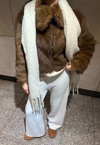 Smeđa lažna krznena kratka jakna s okovratnikom na mašnu, bijeli teksturirani šal, krem hlače širokih nogavica i smeđe gležnjače.