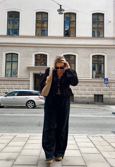 Blusa negra de ojales con mangas abullonadas, con un nudo en el frente, combinada con pantalones negros holgados. Bolso tejido beige, gafas de sol y zapatos de plataforma de madera.