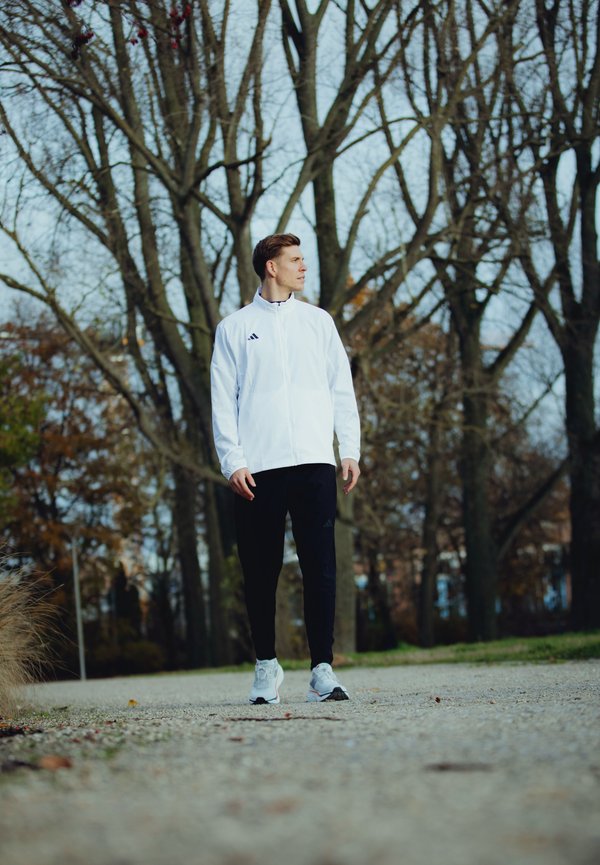Chaqueta blanca con cremallera y acentos negros, pantalones negros y zapatillas deportivas grises; contra un fondo de árboles desnudos y un camino de grava.