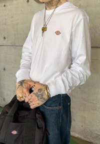 Biela dlhá rukávová tričko s malým, viacfarebným logom Dickies. Čierna taška držená v rukách, denimové nohavice a viacero náramkov na zápästiach.